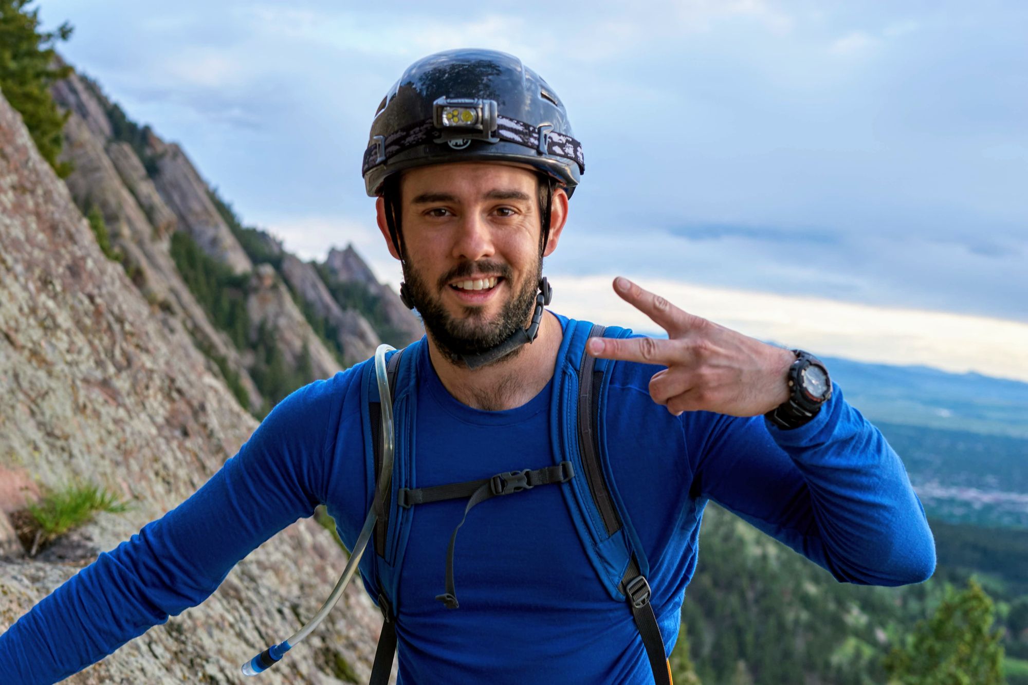 Man in the middle of a rock climbing making the peace sign with his hand. You can see the boulder flatirons in the distance.